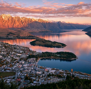 Aerial view of Queenstown, New Zealand at sunset with snowy Remarkables mountains reflecting in Lake Wakatipu.