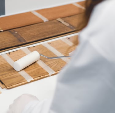 A person in a lab coat uses a small paint roller to apply a clear finish to wood floor samples.