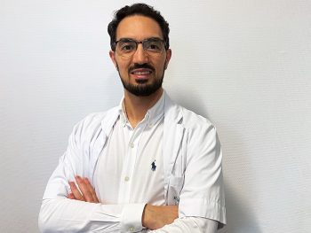 Professional male doctor in white medical coat with arms crossed against a plain background.