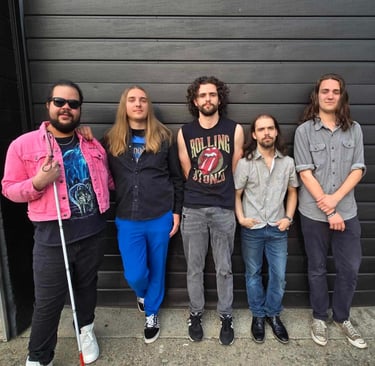 A group photo of five band members standing in front of a wooden and black wall, smiling casually..