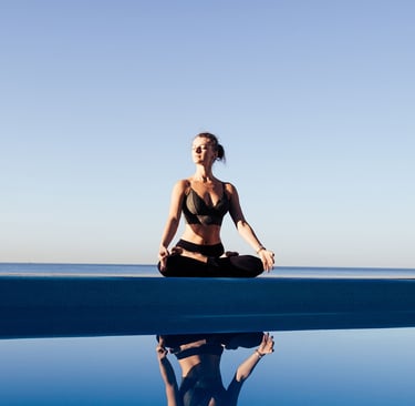 A woman practices yoga meditation in a lotus pose by an infinity pool with an ocean view.