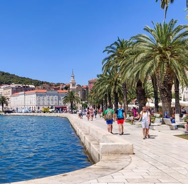 an image of a port with a beautiful blue sea, and a clean boulevard full of people, trees, benches