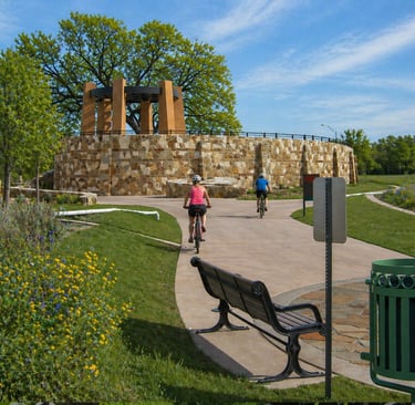 Cyclists ride on a paved trail near a stone pavilion on the San Antonio River Trail.