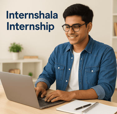 A smiling young South Asian student wearing glasses and a denim shirt works on a laptop at a desk wi