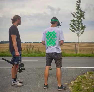two men standing on a road with a camera and a flying fpv drone following a bmw car