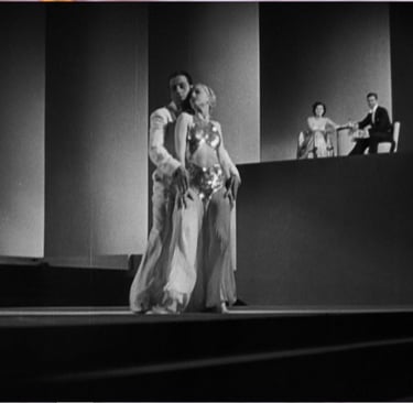 Distant couple in a shadowy nightclub scene watching dancers perform in the moody foreground.