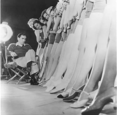 Busby Berkeley seated in a director’s chair, backlit, studying a line of dancers’ legs in perspectiv