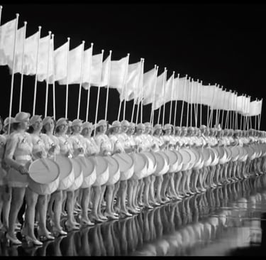 Two rows of chorus girls in short white military costumes with drums and flags in “All’s Fair in Lov