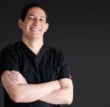 Smiling male healthcare professional in black scrubs with arms crossed against a dark background.