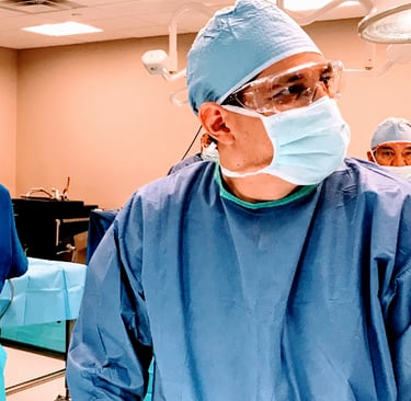 Surgeon in blue sterile scrubs, face mask, and eye protection in an operating room.