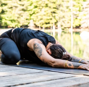 Tattooed woman practicing child's pose yoga on a wooden dock by a calm lake in the forest.