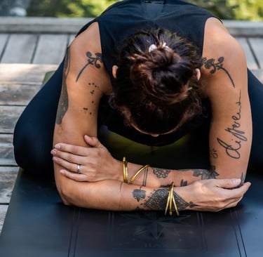 A woman with arm tattoos practicing child's pose on a yoga mat outdoors on a wooden deck.