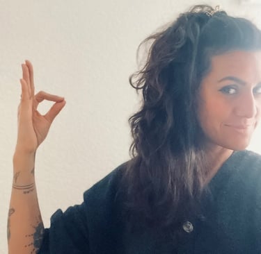 A woman with wavy dark hair and tattoos making an okay hand gesture against a white wall.