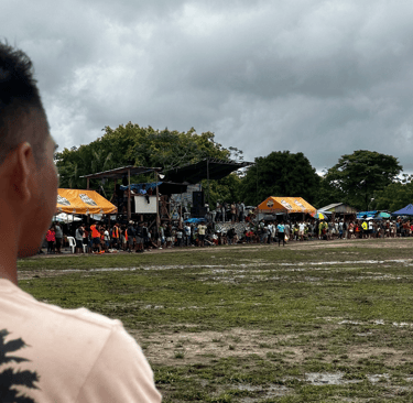 Crowd gathered at a muddy outdoor festival with tents and a stage under a cloudy sky.
