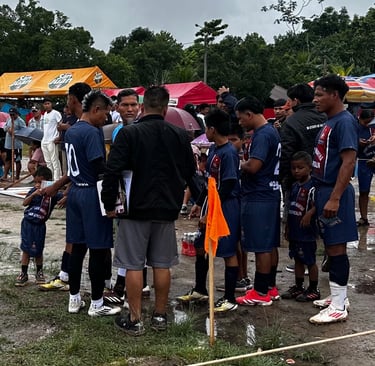 A soccer team in blue uniforms huddles with their coach on a muddy outdoor field during a tournament.