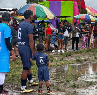 A soccer player and child in matching jerseys stand near a muddy field at a crowded community festival.