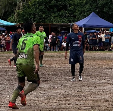 Soccer players competing on a muddy field during a local football match in a rural area.
