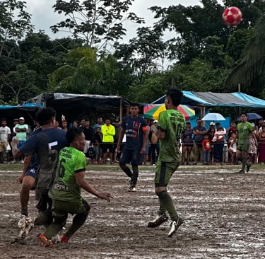 Athletes playing a competitive soccer match on a muddy rural field surrounded by spectators.