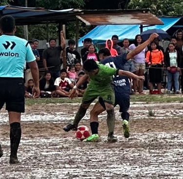 Soccer players competing for a ball on a muddy field during a local match with spectators.