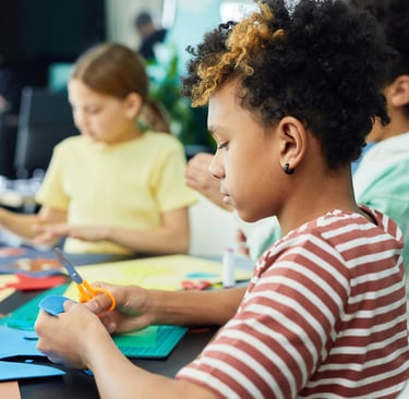 Criança com cabelos cacheados demonstrando alta concentração ao utilizar uma tesoura para recortar papel azul