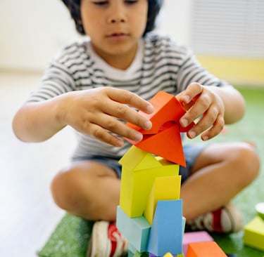 Menino sentado no chão em ambiente terapêutico, demonstrando foco e precisão ao empilhar blocos de montar coloridos.