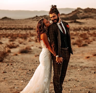 Bride and groom standing together in a desert landscape at sunset, smiling and holding hands