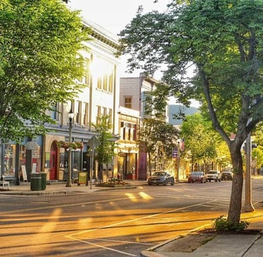 Sunny main street in a historic downtown district with lush green trees and parked cars.
