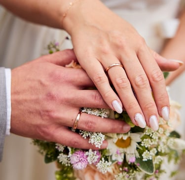 A married couple’s hands with gold wedding rings resting on a floral bridal bouquet.