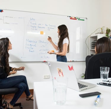 A woman leading a marketing strategy brainstorm on a whiteboard for colleagues in a modern office.