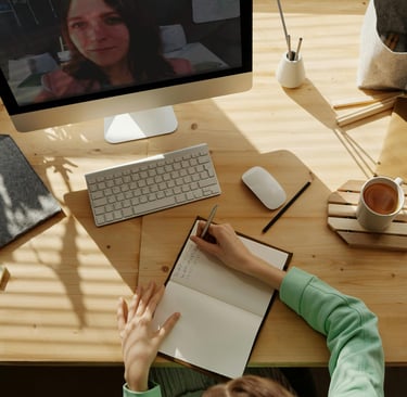 A student taking notes in a notebook during a remote learning video call on a desktop computer.