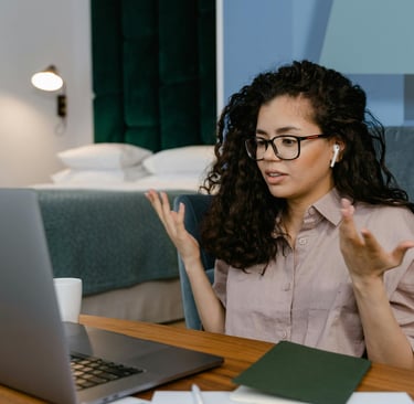 A woman with curly hair wearing glasses joins a video call on her laptop from a home office.