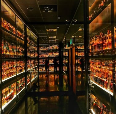 A vast collection of whiskey bottles displayed on illuminated glass shelves in a dark spirits cellar.