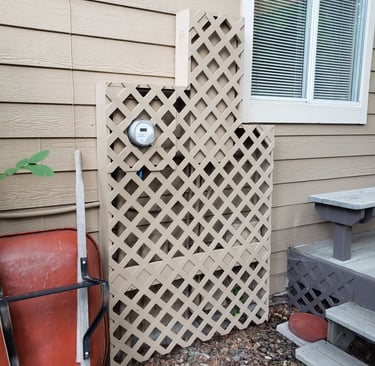Tan garden lattice screen hiding outdoor utility meters on a house exterior next to a red wheelbarrow.