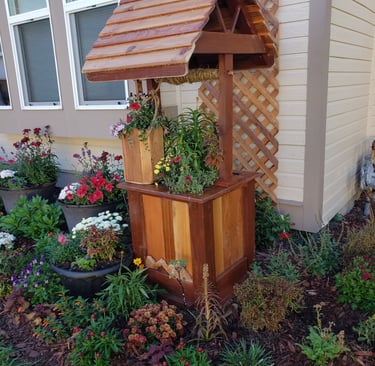 Rustic wooden wishing well planter with a cedar shingle roof in a colorful flower garden.