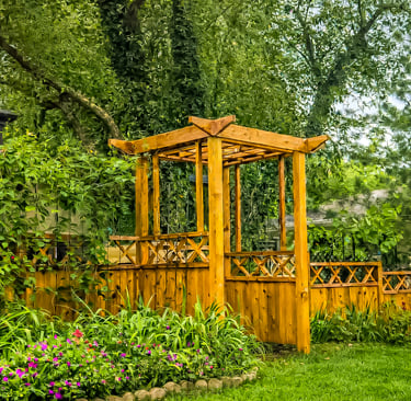 A wooden garden pergola and fence surround a lush backyard with green plants and purple flowers.