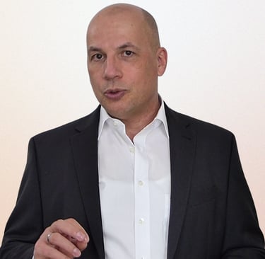 A bald male professional wearing a black suit jacket and white shirt speaking during a presentation.