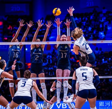 Professional women's volleyball match with players jumping at the net for a block and spike.
