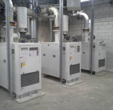 Three industrial air compressor units with ventilation piping in a mechanical room.