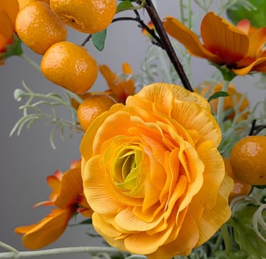 Artificial orange ranunculus flower arrangement with decorative faux citrus fruits and green leaves.