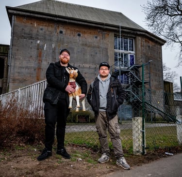 Two men standing outside an industrial bunker building in Hamburg holding a decorative goat statue.