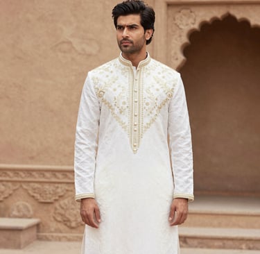 A refined gentleman wearing a terracotta kurta with subtle gold embroidery, standing against a sandstone backdrop.