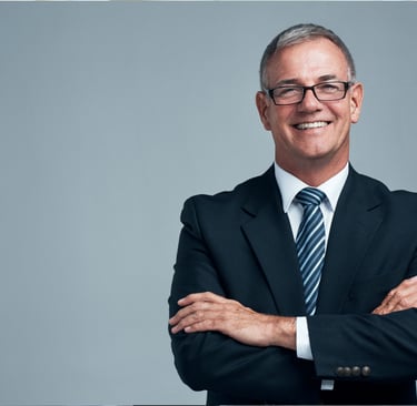 A smiling senior businessman wearing glasses and a suit standing with arms crossed against a grey background.