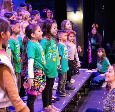 Elementary school children in green holiday choir shirts singing on stage during a winter concert.