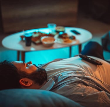 Homme allongé sur un canapé, semblant fatigué, avec une télécommande sur le ventre et des restes de nourriture sur une table