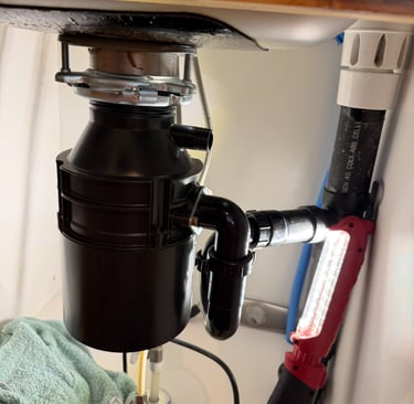 Under-sink view of a new black garbage disposal installation with PVC plumbing and a red work light.