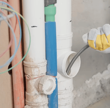 Gloved plumber's hand inserting a drain snake into a PVC pipe to clear a clog in a home's plumbing system.