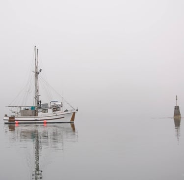 A white commercial fishing boat floats on calm, foggy water near a stone lighthouse beacon.