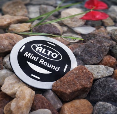 A waterproof Alto Mini Round security tag resting on smooth river stones in shallow water.
