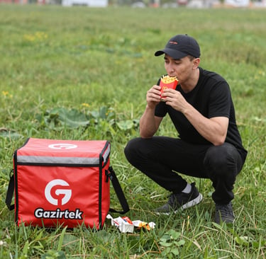 A vibrant delivery driver holding a box filled with fresh ingredients outside a bustling kitchen.