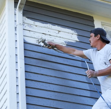 Professional house painter using a power sprayer to spray grey paint to exterior wood siding.